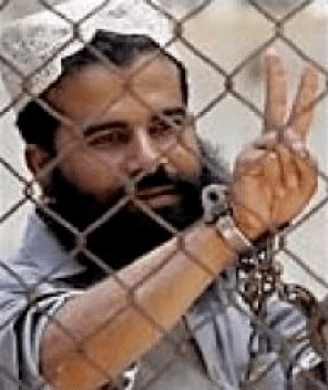 Hassan Ghul doing a peace sign inside prison with handcuffs and sporting a thick beard while wearing gray clothes and white hat.