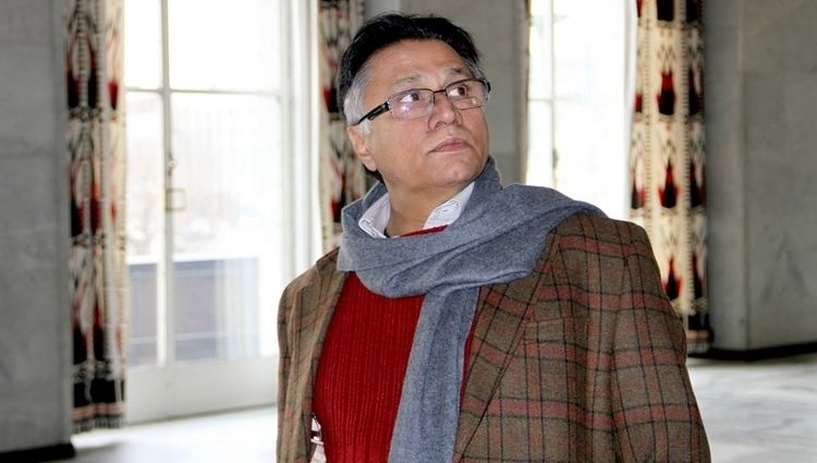 Hassan Nisar wearing eyeglasses, a checkered coat, a red shirt, and a gray scarf.