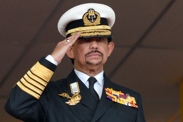 Hassanal Bolkiah with a serious face while doing a salute, with a beard and mustache and a ring on his right finger ring, wearing a multi-colored forage cap, and a black and yellow service uniform with special skills and identification badges over white long sleeves, and a black necktie.