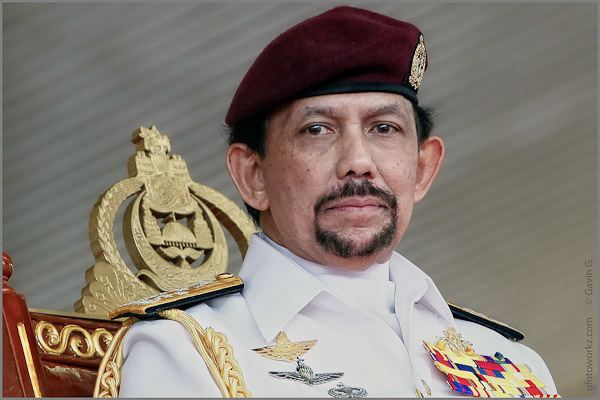 Hassanal Bolkiah with a serious face while sitting on a throne, with a beard and mustache, wearing a maroon forage cap with a badge on it, and identification badges in a white service uniform