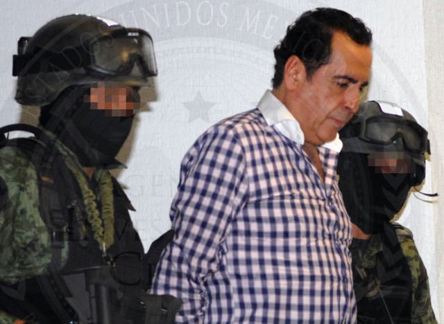 Héctor Beltrán Leyva with a sad face and wearing a checkered white and blue polo shirt while arrested by two policemen.