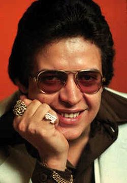 Héctor Lavoe smiling with his right hand on his cheek and wearing a brown shirt and white coat with eyeglasses and gold jewelries