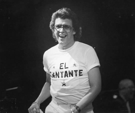 Héctor Lavoe laughing in his long hair and wearing a white shirt and white pants with eyeglasses