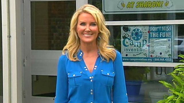 Heather Childers smiling, with wavy blonde hair and white painted glass door and panel window in the background with writings of "Camp invention" and "DON'T MISS THE FUN!" on a blue and white banner and a plant on the left. She is wearing a blue long sleeve top.