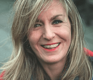 Heather McCartney with a big smile and blonde hair while wearing earrings and a blue blouse under a red blazer