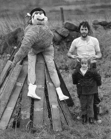 Paul McCartney and Heather McCartney are standing near the pieces of wood that formed into a triangle with a teddy bear on the top of it wearing a hat, sunglasses, jacket, and pants. Paul is wearing a white t-shirt and pants while Heather is wearing shoes, pants, and a blouse under the jacket