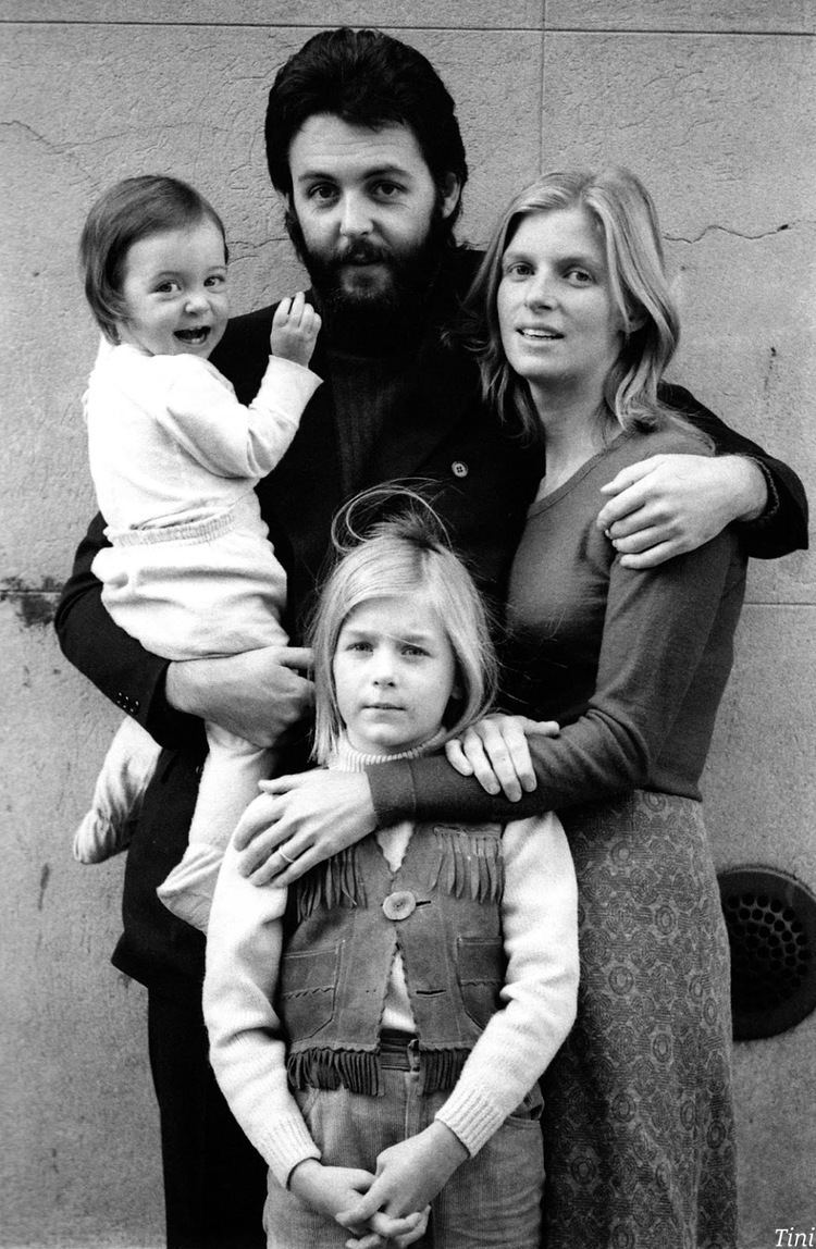Mary is smiling (left) while being carried by Paul with a mustache and beard (center) beside him is Linda (right) and in front of them is Heather McCartney with a serious face (front center). Mary is wearing a long sleeve shirt and pajama, Paul is wearing pants and a shirt under a black coat, Linda is wearing a long sleeve blouse and skirt while Heather is wearing a long sleeve blouse under a vest