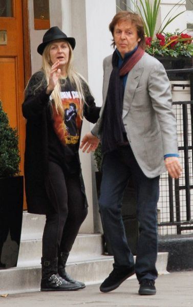 Heather McCartney with a shocked face while Paul McCartney is looking at something. Heather is wearing a black hat, black blazer, black printed shirt, black pants, and shoes while Paul is wearing denim pants, black shoes, and a blue long sleeve under a gray coat, and maroon scarf