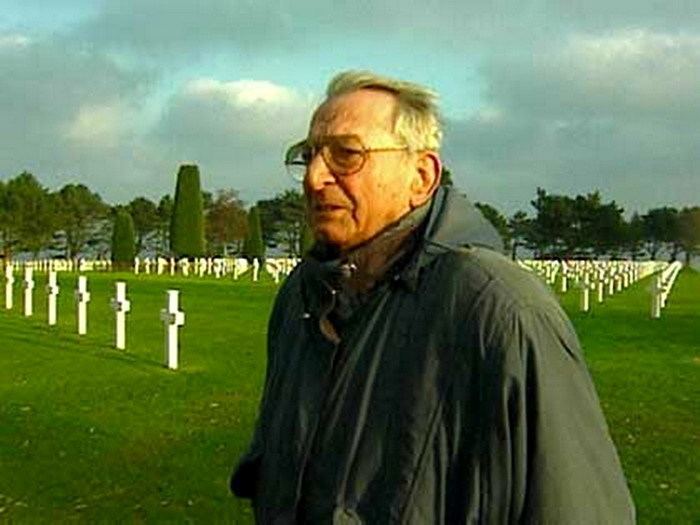 Heinrich Severloh was saddened looking from afar in a cemetery with multiple crosses in the background, he has white hair wearing an eyeglass, and a gray polo with a gray necktie under a black hoody jacket