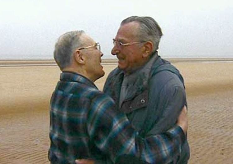 Heinrich Severloh (right) and David Silva (left) hugging and smiling at each other in Omaha Beach. Heinrich has white hair, wearing an eyeglass, and a gray polo with a gray necktie under a black hoody jacket while David has white hair too and wearing eyeglass and a checkered long sleeve