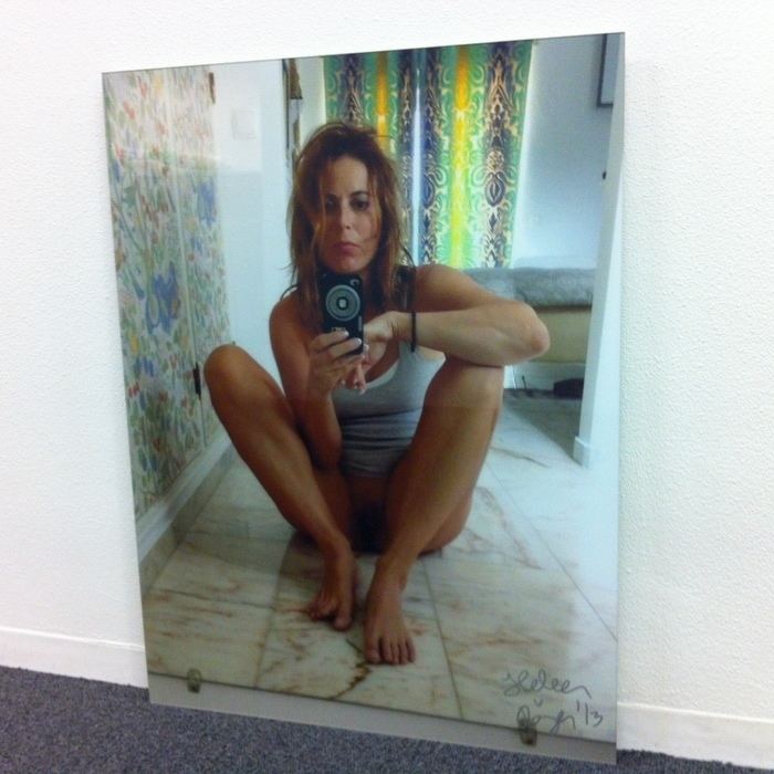 Heleen Royen is serious, has brown hair, vagina and pubic hair is seen, sitting down on the floor inside a room, and has colored green, blue, and yellow curtains behind, on her right is a colored wallpaper with a design, right hand holding a camera, left hand on her knee, wearing a white-gray sleeveless top, naked on the bottom.