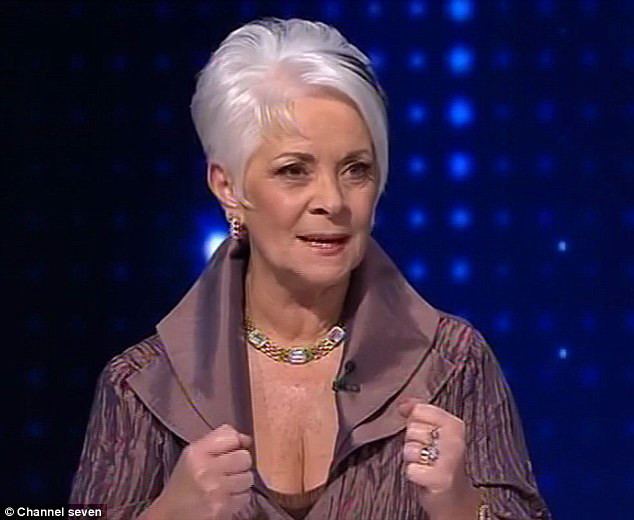 Helen Richey smiling, with clenched fists, white hair, wearing earrings, a necklace, rings, and a brown top showing her cleavage with a wireless microphone on it.