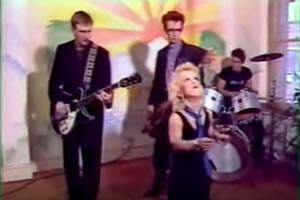Helen Wellington Lloyd dancing while clapping both her hands together with a band in the background, a three-man holding band instruments, 2 men are holding a guitar wearing a black long sleeves and black pants and the other one was playing as a drummer, wearing a black shirt.  Helen has short blonde hair, wearing a watch on her left wrist, a black sleeveless dress, and a purple scarf around her neck