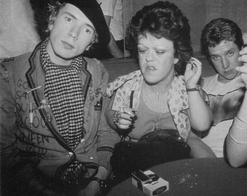 (From left to right) Johnny Rotten, Helen Wellington Lloyd, and a man are sitting together on a couch with a cigarette box on the table in front in a black and white picture. Johnny is seriously looking, has black hair and wearing a  black hat, a watch on his left wrist, striped polo under a coat that has many pins on it while Helen, is looking below with her left hand holding her hair and a black bag on her lap, she has a short hair, wearing a necklace, rings on her right fingers, a watch on her left wrist, and a shirt under a polka dots blazer