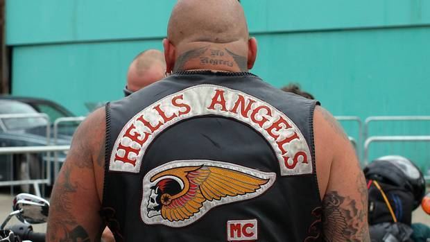 A Hells Angels member with a bike & helmet in the background, he has a bald head and tattoos on his body is wearing an earring, black sleeveless jacket with Hells Angels' logo on the back