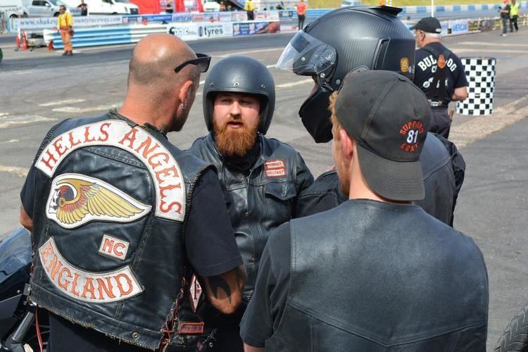 Four men talking to each other. The man (1st from left) with a bald head is wearing an earring, a black shirt under a black sleeveless jacket with the Hells Angels logo. The man (2nd from left) with a mustache and beard is wearing a black leather jacket and black helmet. The two other man is wearing a black helmet, cap, and black t-shirt under a black sleeveless jacket