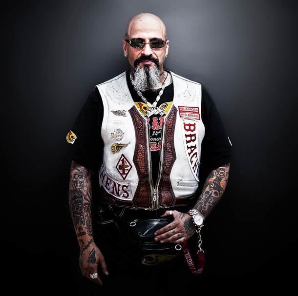 A Hells Angels member with a bald head, tattoos on his body, mustache, beard, and hand on his belt bag while wearing sunglasses, necklace, wristwatch, ring, and a black printed t-shirt under white sleeveless jacket with patches