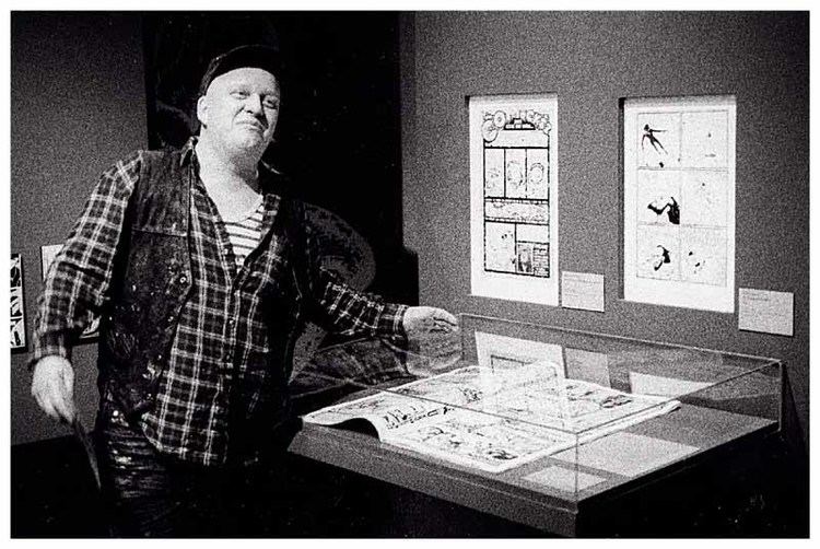 Henriette Valium posing beside the comic book while wearing checkered long sleeves, vest, and cap