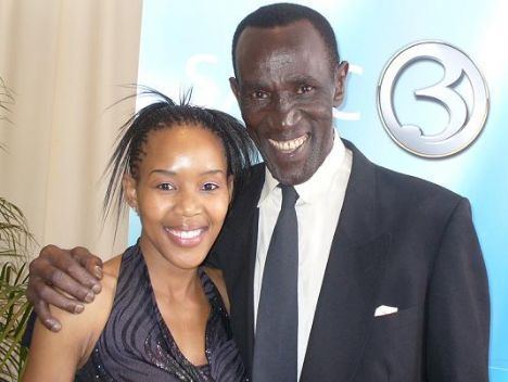 Henry Cele smiling while wearing a black coat, white long sleeves, and gray necktie and the woman beside him wearing a gray and black blouse