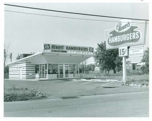 Henry's Hamburgers from 1974 a Vintage Buffalo in New York, in a road side, has white building with glass window walls, with store name at the top, “15 Henry's Hamburgers 15” with a field with grass and trees and a parking lot with large road sign of Henry’s Hamburgers.