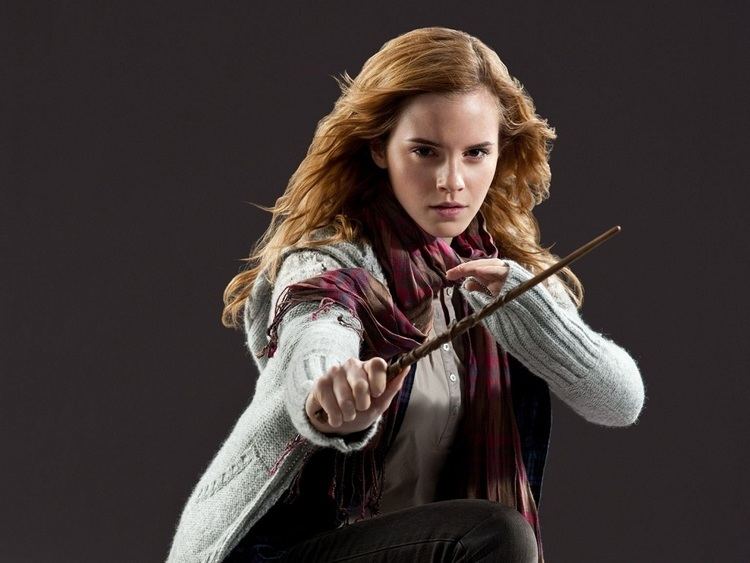 Emma Watson as Hermione Granger with a serious face, standing, holding her long, made of vine wood, and possessed a dragon heartstring core wand with her other hand in front of her face and hair blown by air, in the J.K. Rowling novel series, Harry Potter. Emma with curly blonde hair wearing a scarf and light brown polo under a grey knitted hoodie