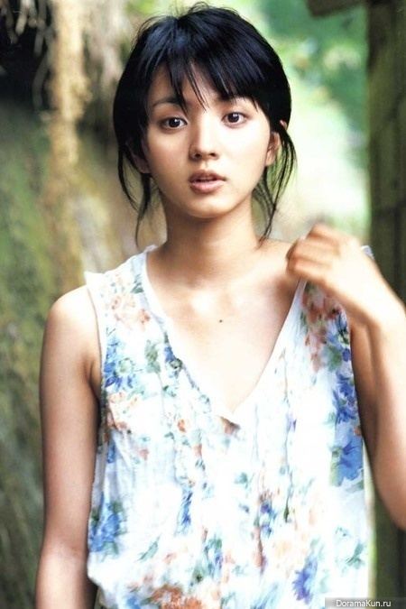 Hikari Mitsushima is smiling, has black hair, her left hand on her left shoulder, a mole on her chest, wearing a white with blue and red floral sleeveless top.