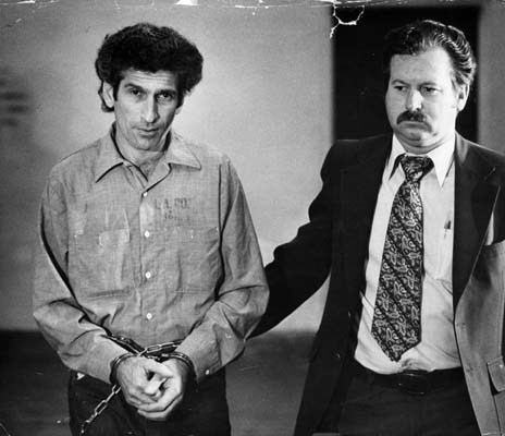 Angelo Buono with handcuffs and wearing long sleeves and pants while the man beside him wearing a coat, long sleeves, and necktie