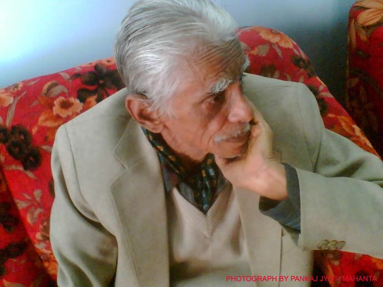 Hiren Bhattacharyya sitting on a couch wearing a brown and gray shirt, and a brown coat