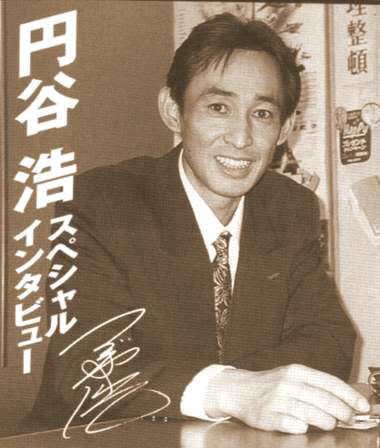 Hiroshi Tsuburaya smiling while wearing a black coat, white long sleeves, and necktie