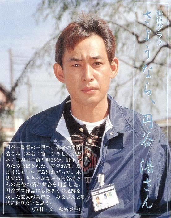 Hiroshi Tsuburaya with a furrowed brow while wearing a blue jacket, brown sweatshirt, white shirt, and identification card