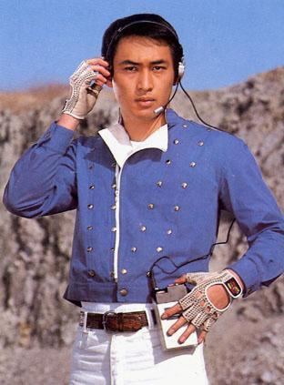 Hiroshi Tsuburaya holding his headphone while wearing silver gloves, blue long sleeves with gold buttons, brown belt, and white pants