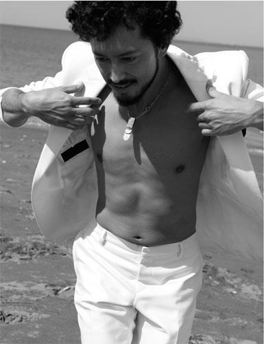 Hiroyuki Ikeuchi is smiling while looking on the ground and holding his white coat, with curly hair, and wearing white pants.