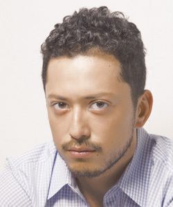 Hiroyuki Ikeuchi with a serious face, curly hair, with beard and mustache, and wearing blue striped long sleeves.