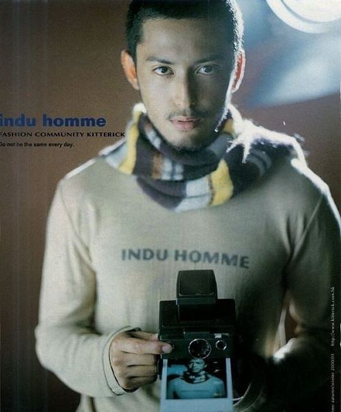 Hiroyuki Ikeuchi with a serious face while holding a camera with his photo, wearing a brown long sleeves shirt and checkered scarf.
