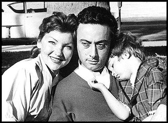 Honey Bruce is smiling while leaning at Lenny Bruce with a serious face while carrying their son Kitty Bruce while sleeping. Honey wearing a blouse, Lenny is wearing a sweatshirt while Kitty is wearing a checkered polo shirt.