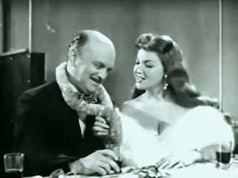 Honey Bruce smiling while looking at a bald man. Honey with wavy long hair and wearing a white fur dress with a plunging neckline while the man is wearing a black coat.