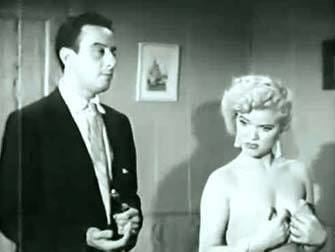 Honey Bruce with a sad face beside a man with a serious face. Honey is wearing earrings and a tube dress while the man is wearing a black coat over white long sleeves and a tie.
