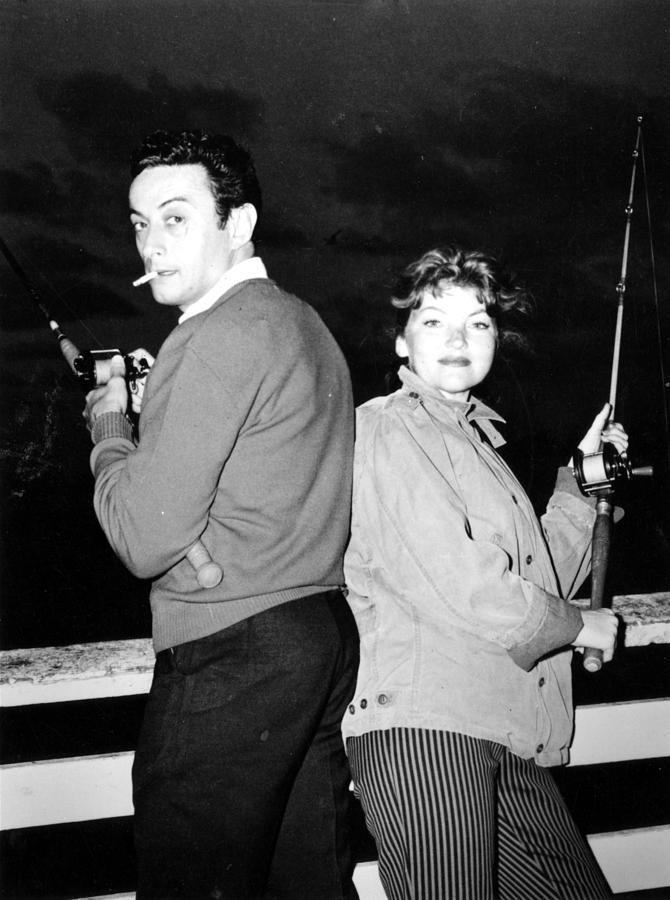 Honey Bruce with a tight-lipped smile while Lenny Bruce is smoking and both are holding fishing rods. Honey wearing a jacket and black striped pants while Lenny is wearing a sweatshirt and black pants.