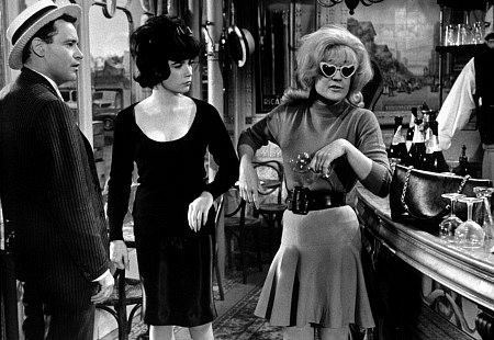 Jack Lemmon and Shirley MacLaine are looking at Hope Holiday while Hope is holding a wine glass with a serious face. Jack is wearing a hat, pants, and a long sleeve under a coat. Shirley is wearing a long sleeve dress. Hope is wearing sunglasses with a heart shape, long sleeve blouse, and a skirt with a belt in a scene from the 1963 American romantic comedy film, Irma la Douce
