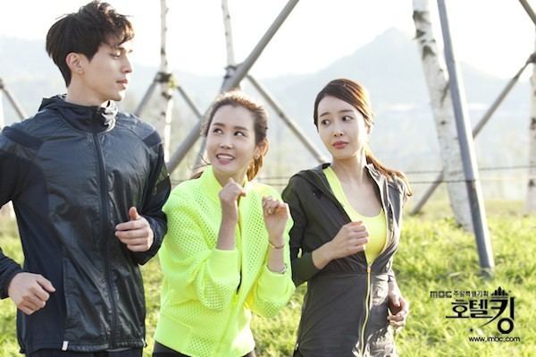 In the movie scene Hotel King 2014, Lee Dong Wook is serious, running, with both hands in a running pose, has brown hair, wearing a black jacket and pants, in the middle is Kim Gyu Sun smiling, looking at Lee Dong Wook, running, while her hands in a boxing position, has long brown hair wearing a yellow jacket and a  bracelet on her left hand, and black pants, at the right is Lee Da Hae serious, looking at Kim Gyu Sun while running, has brown long hair wearing a yellow tank top and a gray jacket.