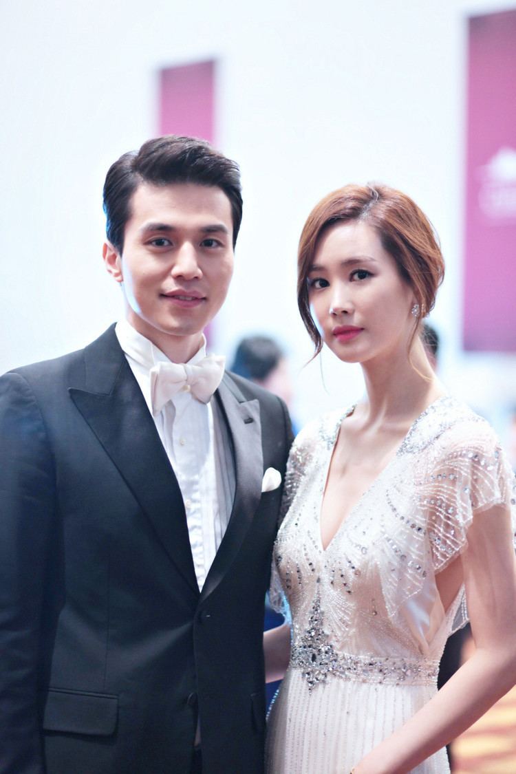 On the left Lee Dong Wook is smiling, standing hands down, has black hair, wearing white long sleeves with a white bow tie and black coat, at the right is Lee Da Hae serious, standing, right arm behind Lee Dong Wook's waist has brown hair wearing silver earrings, a white dress with silver pattern beads.