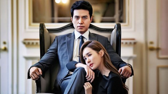 In the movie scene Hotel King 2014, Lee Dong Wook is serious, sitting in a black and gray chair, both hands resting on the arm top of the chair, in a white room hotel, has black hair wearing white long sleeves, a black necktie, black pants, and black coat, at the right Lee Da Hae is serious, sitting on the floor, right arm on Lee Dong Wook legs, and left hand below the knee, has long brown hair, wearing a black dress.