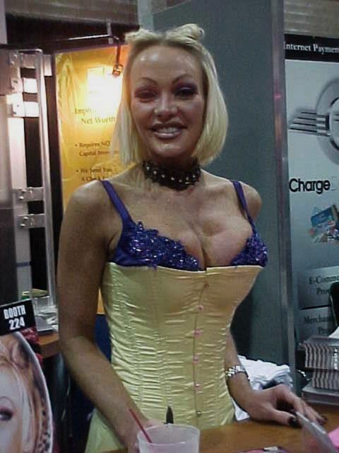 Houston smiling, with short blonde hair, wearing a black choker, and a yellow and purple spaghetti top.