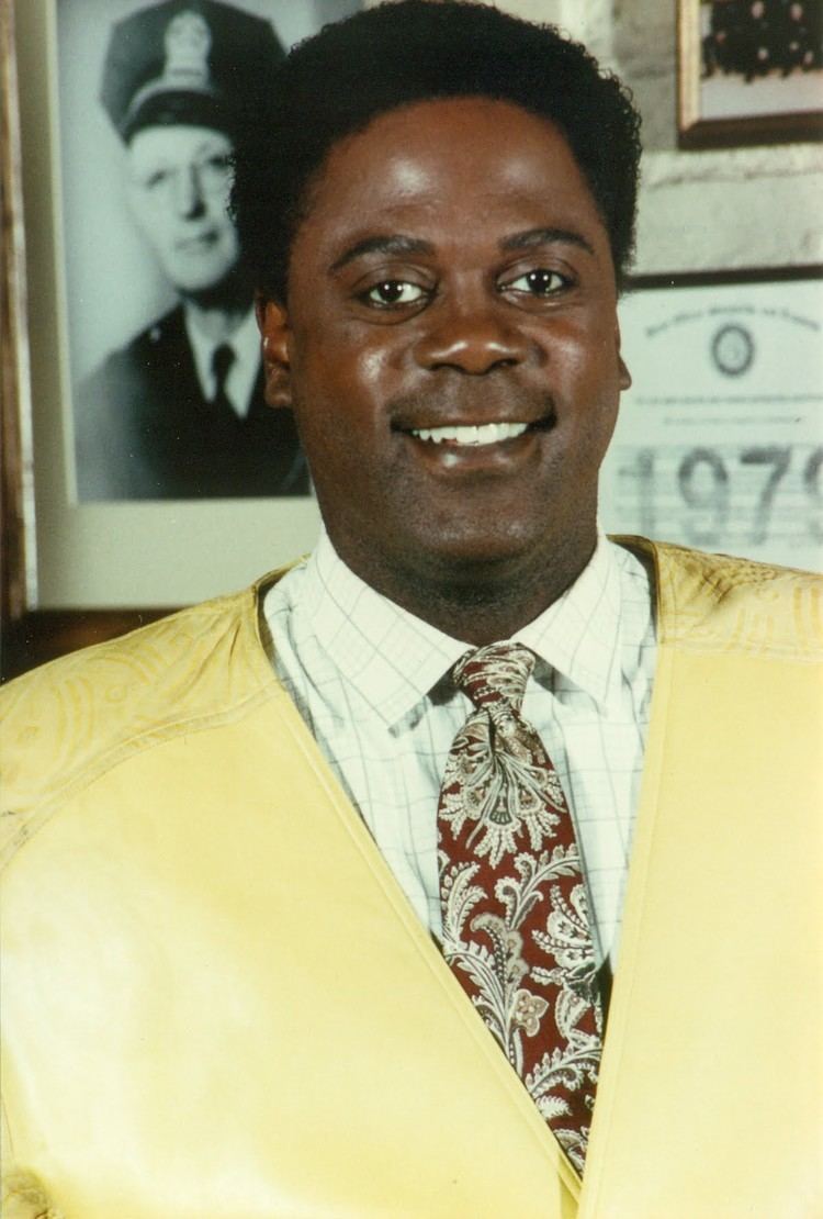 Howard Rollins smiling and wearing yellow vest, white checkered long sleeves and neck tie