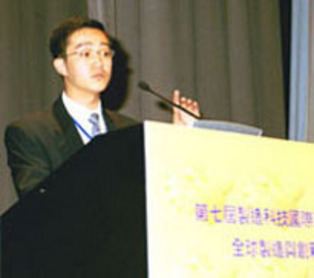 In a conference room with a large brown curtains and speaker's table with mic on top and a chinese characters in front, Hu Haifeng is serious, eyebrows up, standing, holding a mic with his left hand, has black hair wearing eyeglasses, a white polo with blue necktie under a black coat,