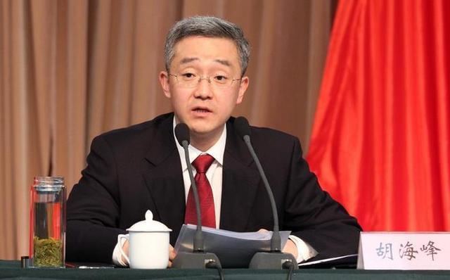 In a conference room with a large brown and red curtains and black table with bottle of water a tea cup with cover a mic and a name plate with chinese characters on top, Hu Haifeng is serious, eyebrows up, sitting, speaking through the mic, holding a paper with his hands, has white hair wearing eyeglasses, a white polo with red necktie under a black coat,