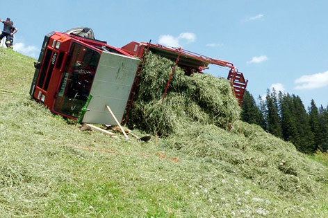 A red and white farm truck filled with grass fells down on the hill with two people standing in the background.