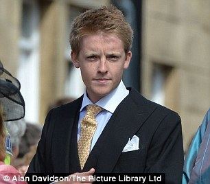 Hugh Grosvenor posing while wearing a black suit and a sky blue long sleeve and a yellow tie.