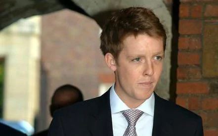 Hugh Grosvenor looking at something while wearing a black formal attire with a gray tie.