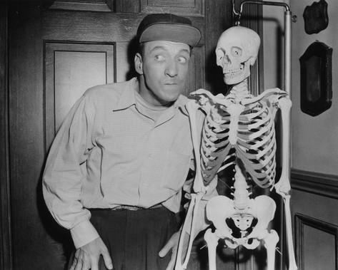 Huntz Hall looking at the left while standing beside the skeleton and wearing a cap, long sleeve, and pants